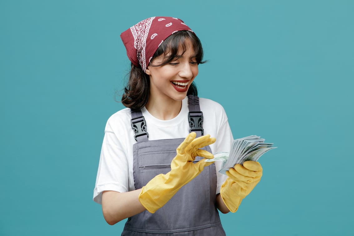 joyful-young-female-cleaner-wearing-uniform-bandana-rubber-gloves-holding-money-looking-it-counting-money-isolated-blue-background